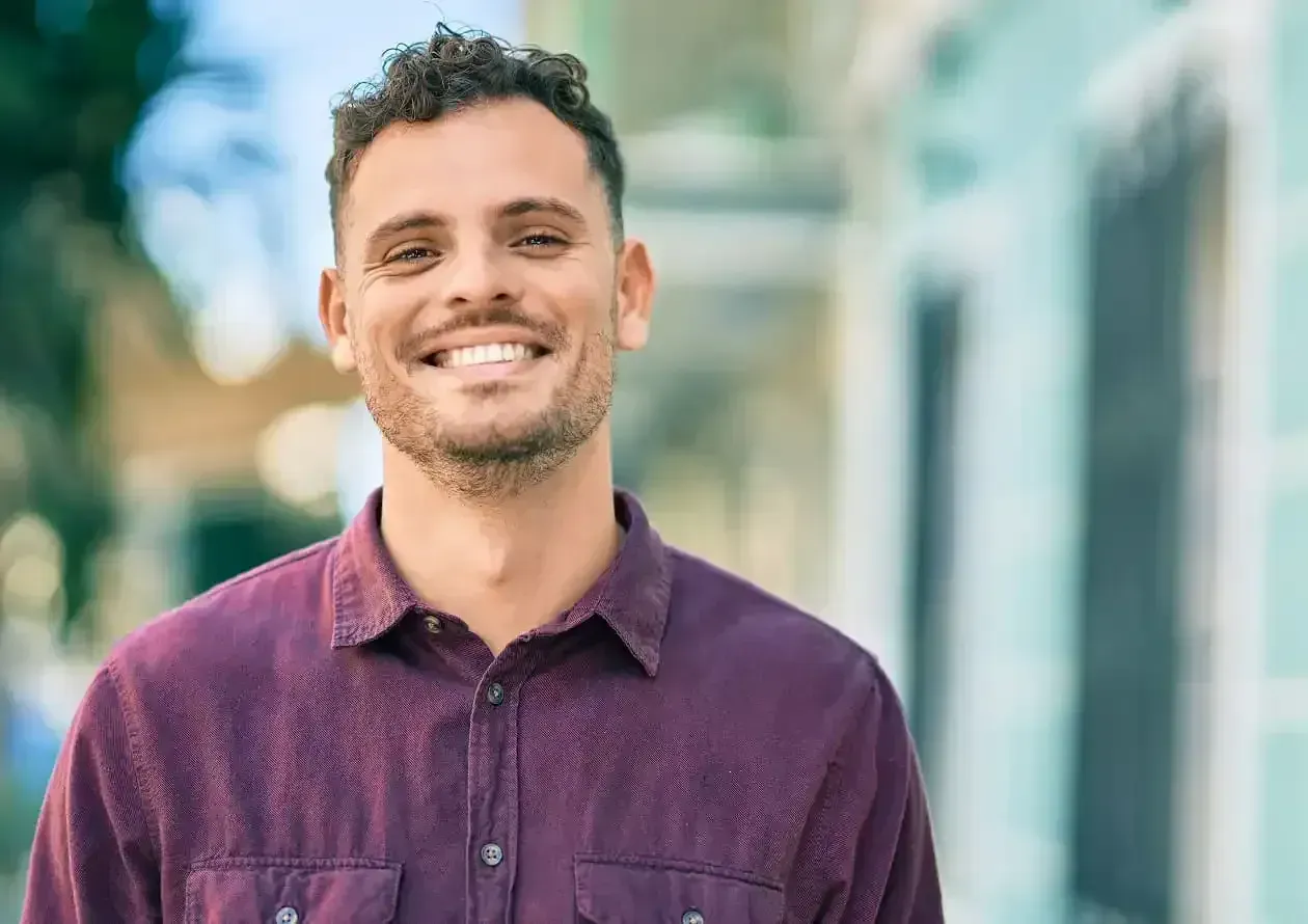 Smiling man with curly hair and beard wearing a maroon button-up shirt outdoors.